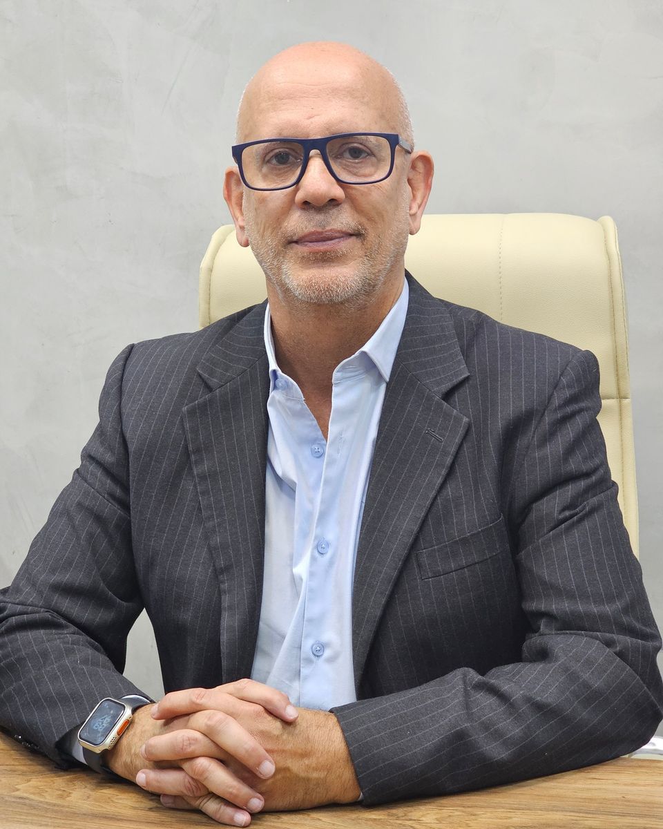 Retrato do médico sentado a uma mesa de madeira clara, com as mãos entrelaçadas apoiadas sobre o tampo. Veste terno cinza escuro de risca de giz e camisa azul clara, com óculos de armação azul marinho. O fundo é uma parede cinza clara e lisa.