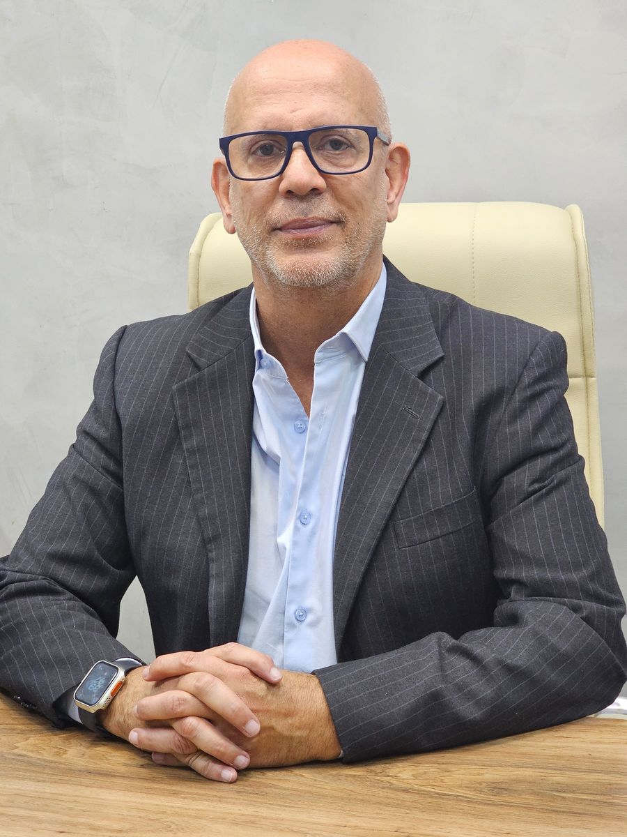 Retrato do médico sentado a uma mesa de madeira clara, com as mãos entrelaçadas apoiadas sobre o tampo. Veste terno cinza escuro de risca de giz e camisa azul clara, com óculos de armação azul marinho. O fundo é uma parede cinza clara e lisa.